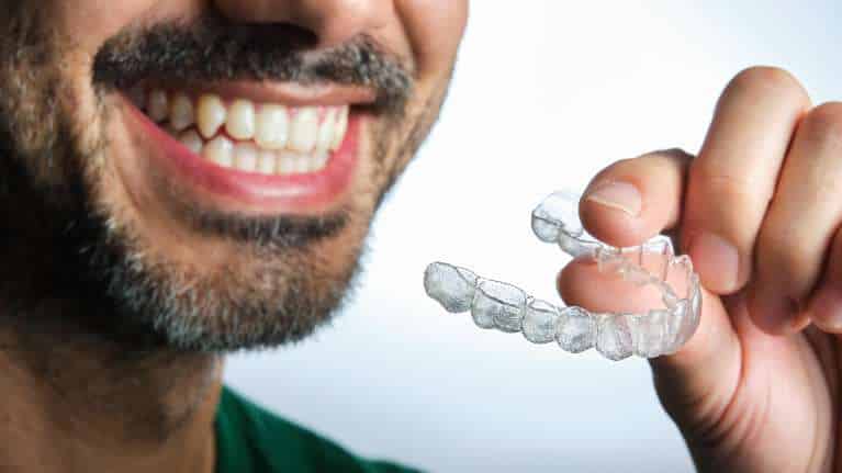 A young man with a confident smile holds a clear Invisalign aligner tray in his hand
